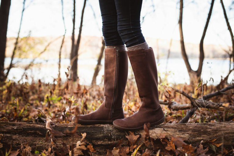 A close up of a woman walking on a log in the woods while wearing Vivobarefoot Ryder boots, a popular tall leather barefoot boot.