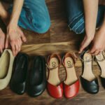 A row of pretty stylish women's barefoot dress shoes in a row on a wood floor with the arms of two people showing as they reach for the shoes
