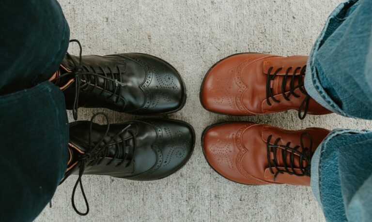 Top down view of 2 people wearing Zaqq Barefoot wide toe box leather boots with Broque detailing. They are standing toe to toe. The right pair is brown and the left is black