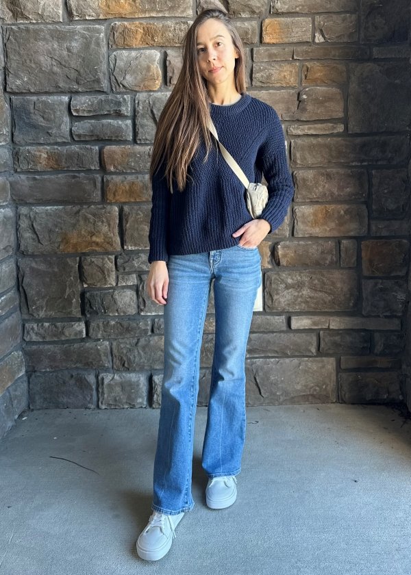 A full body photo of a woman in flare leg jeans and a navy blue sweater looking at the camera. She has Splay Rev LT sneakers with an anatomical toe box on her feet.