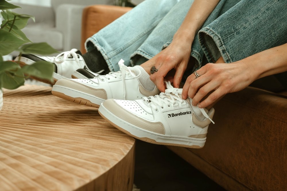 Two pairs of legs with their sneakers resting on the edge of a wooden table. They are both wearing Barebarics Wave in white leather with neutral tan or black accents